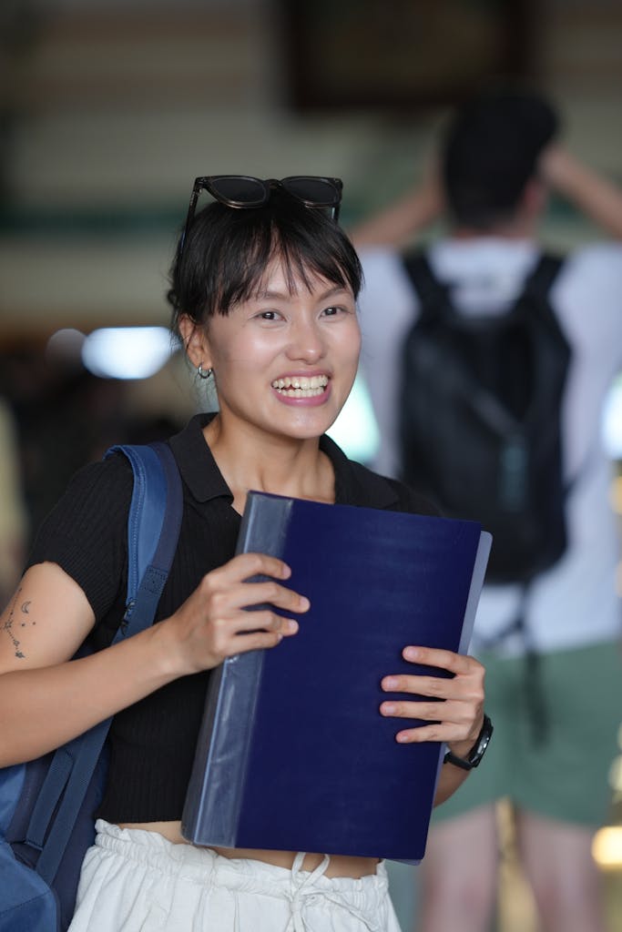 Smiling young adult holding a notebook in an indoor setting, expressing happiness.