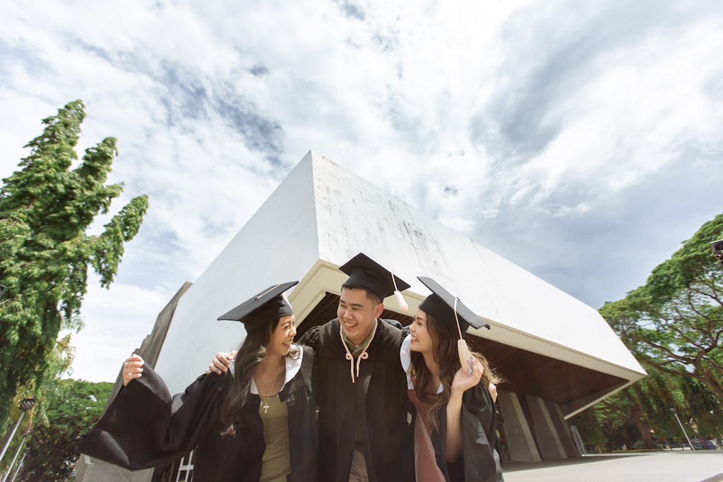 Three graduates in caps and gowns celebrating together outdoors near a modern building.